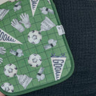 A close-up of a green soccer-themed fabric item, likely a pouch or mat, featuring a grid pattern with soccer balls, gloves, whistle, and pennant flags printed with 'GOAAL!'. The fabric is laid flat on a dark textured surface.