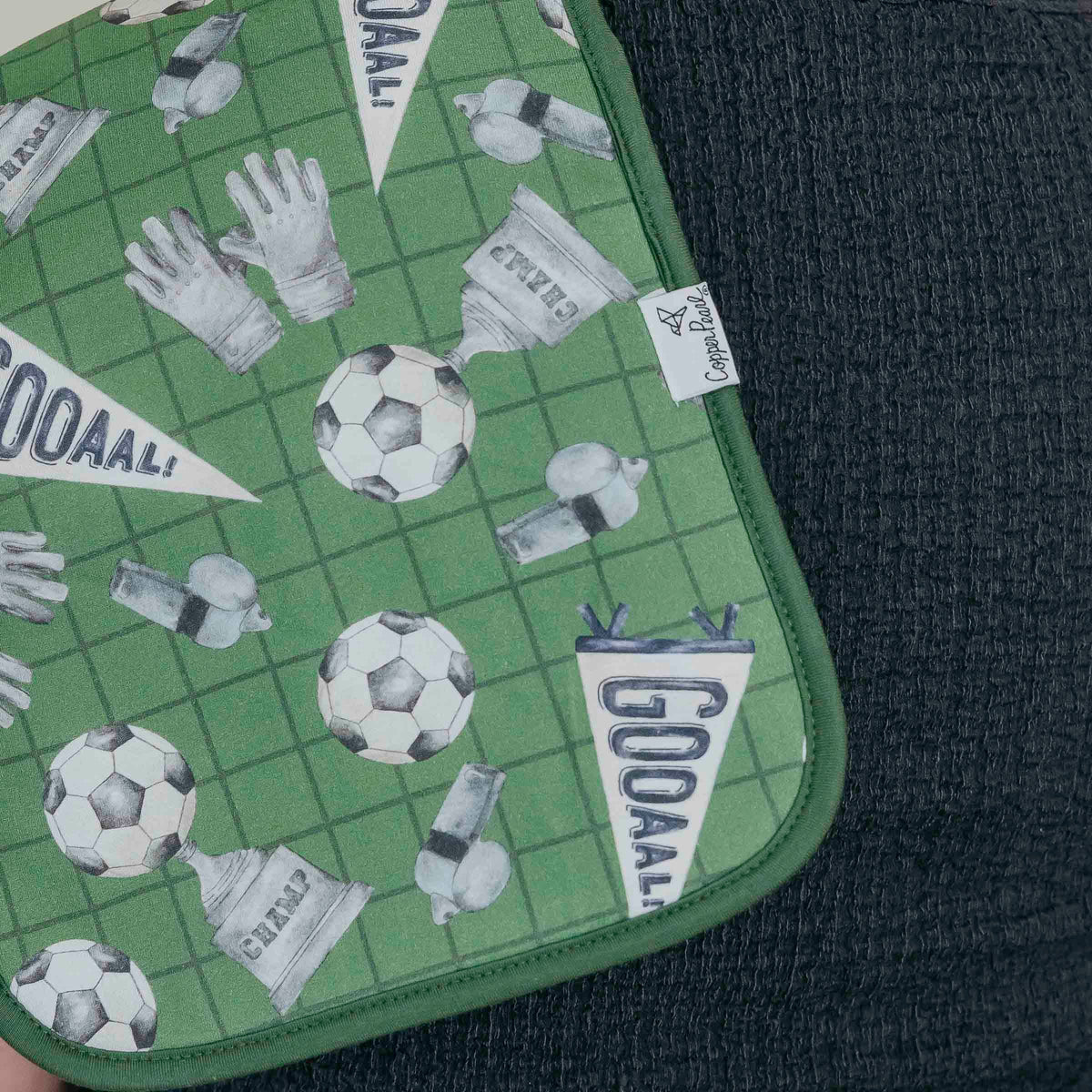 A close-up of a green soccer-themed fabric item, likely a pouch or mat, featuring a grid pattern with soccer balls, gloves, whistle, and pennant flags printed with 'GOAAL!'. The fabric is laid flat on a dark textured surface.