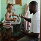 A father watching and smiling as his young child in a striped shirt plays with toy whales at a bathroom sink in a green-tiled room.