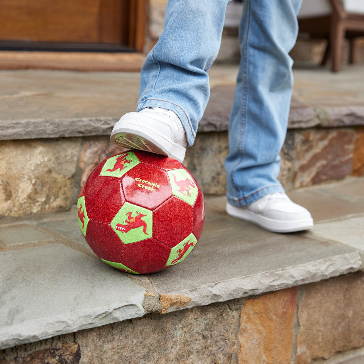 Red soccer ball with green patterns on a stone step, person wearing blue jeans and white shoes.
