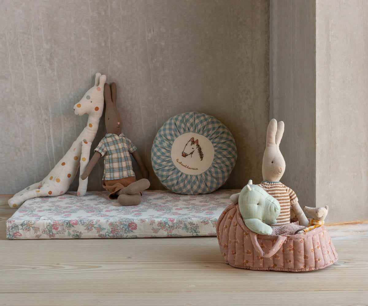 Children's toys including a giraffe, rabbit, and other plush items on a floral mattress against a neutral wall.