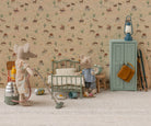 Two Maileg mouse toys in a miniature room setting with patterned wallpaper and carpet; one mouse on the left operates a toy vacuum cleaner next to a thermos and canister, while another mouse on the right stands beside a mint green single bed and a matching storage cabinet with a blue lantern on top.