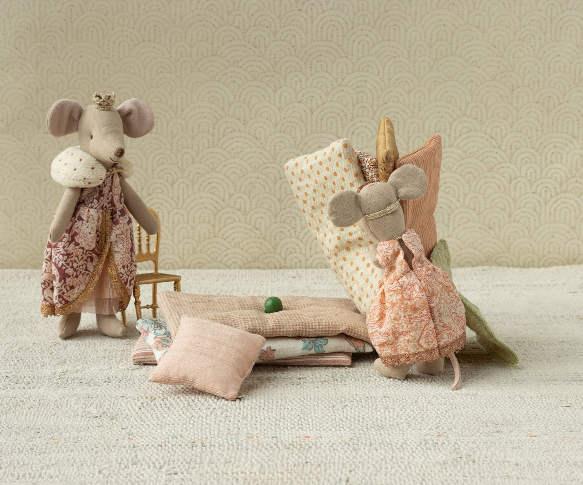 A Maileg toy set showing a Big Sister mouse standing next to a stack of patterned mattresses, a miniature pillow, and a tiny green pea resting on the bed setup.