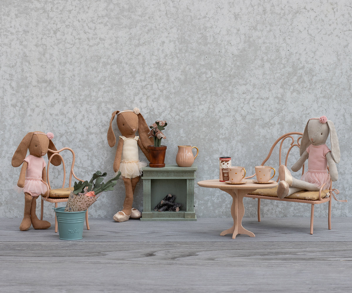 Two Maileg toy rabbits are sitting in a miniature room with a table, chairs, a small fireplace, and decorative accessories including potted plants and a crate of carrots.