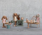 Two Maileg toy rabbits are sitting in a miniature room with a table, chairs, a small fireplace, and decorative accessories including potted plants and a crate of carrots.
