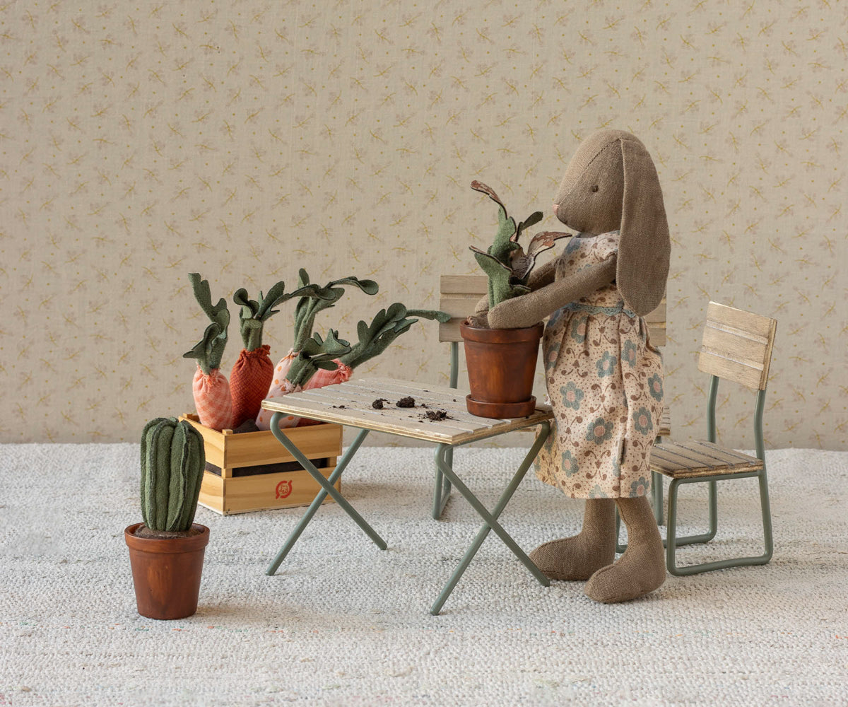 A Maileg toy rabbit wearing a floral dress stands by a miniature table and chairs, holding a small potted plant. Other miniature potted plants, carrots in a crate, and a cactus sit nearby in a cozy room setting with wallpaper.