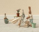 Three Maileg toy rabbits are gathered in a miniature room with a table, chairs, a broom, a bucket, and a crate of carrots, creating a playful scene against patterned wallpaper.