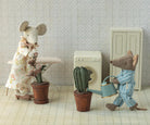A Maileg toy mouse in striped pajamas holding a blue watering can walks toward a potted cactus, while another Maileg mouse in a floral dress stands by a table holding a small potted plant in a miniature room with patterned wallpaper.