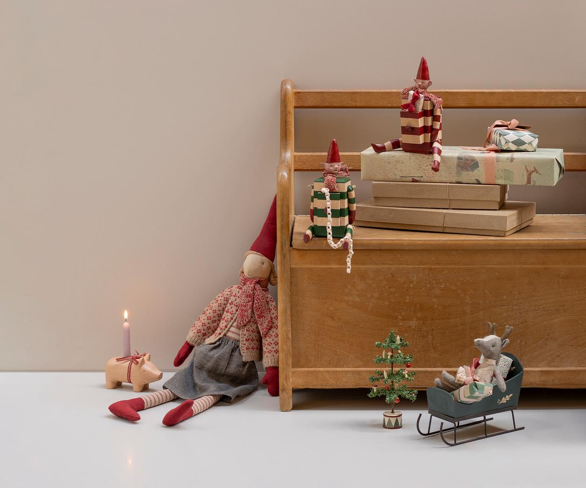 A miniature green wooden Maileg toy mouse sleigh on a wooden floor, with a larger plush toy duck, gnome dolls, a small suitcase, and holiday decor arranged in a rustic room setting.