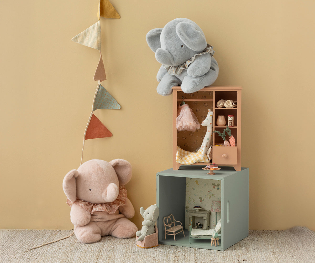 Two plush elephants and a small dollhouse on a beige floor with a decorative wall hanging.