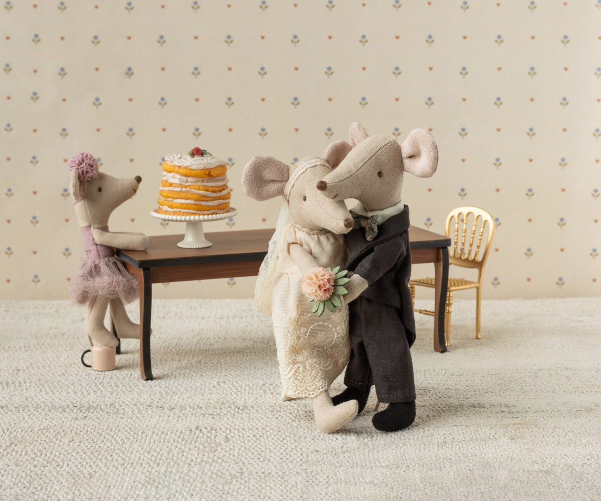 Two toy mice in wedding attire standing next to a cake on a small table with decorative wallpaper in the background.