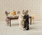 Two toy mice in wedding attire standing next to a cake on a small table with decorative wallpaper in the background.