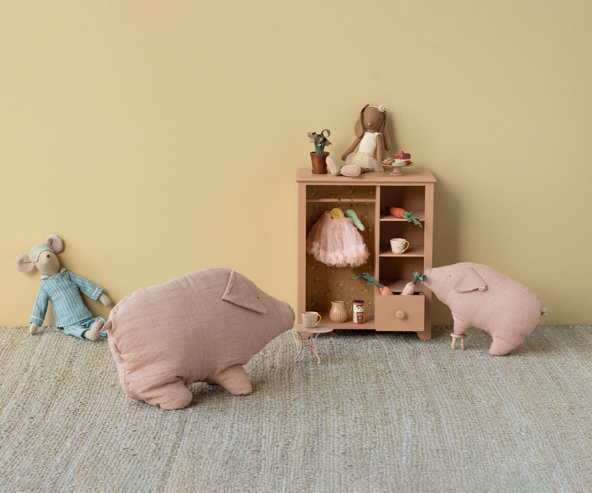 A soft, dusty pink linen stuffed pig toy standing next to a miniature wooden wardrobe on a textured rug, alongside a plush mouse and a larger pillow-like pig.
