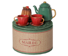 A Maileg Christmas tea party set featuring a round decorative gift tin, a green metal teapot with a gold knob and handle, two red teacups and saucers, and a miniature cake on a scalloped plate.