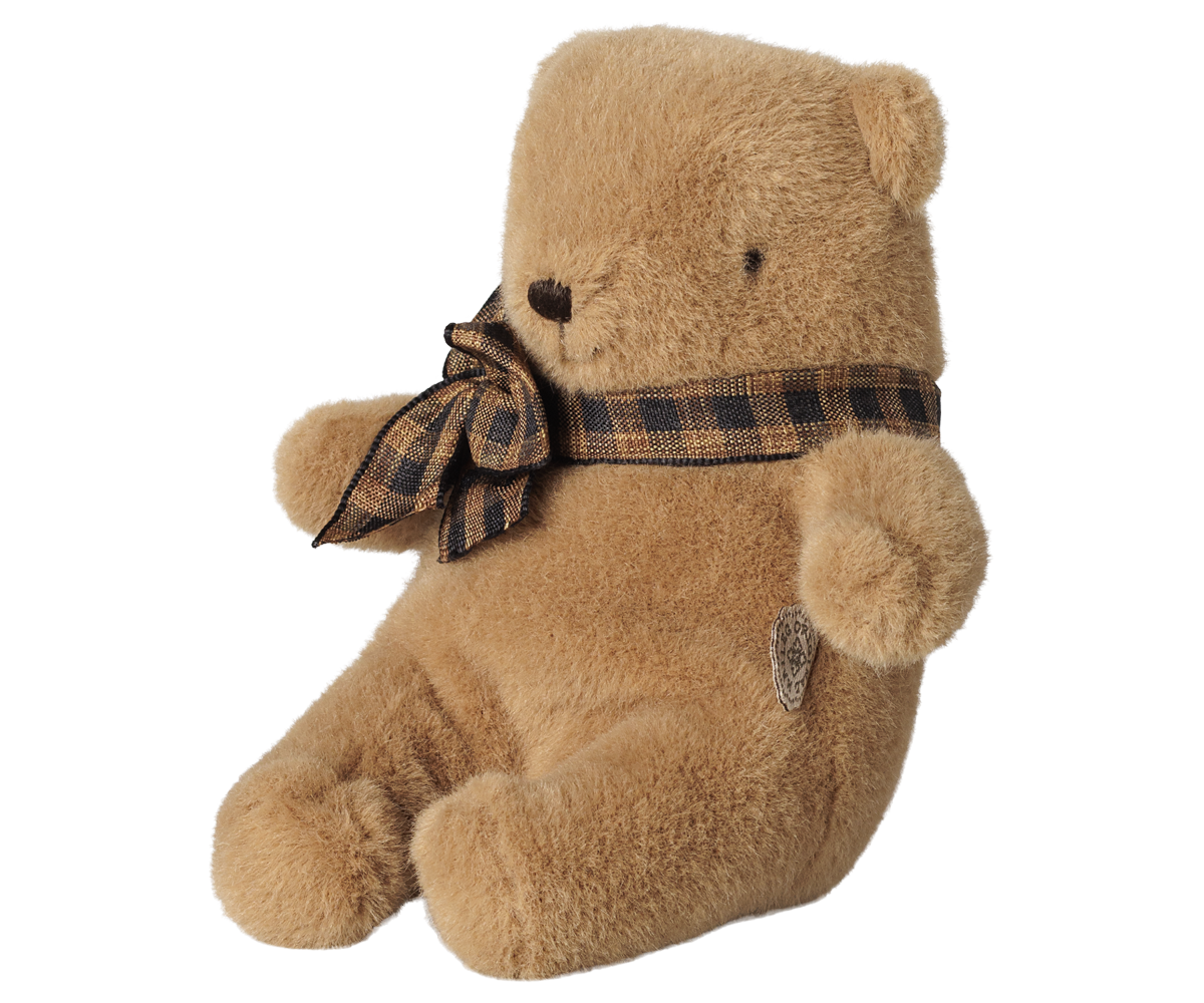 A brown Maileg mini teddy bear sitting with its legs extended forward, wearing a brown and black plaid bow around its neck.