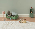 A styled children's bedroom scene with a green wooden bed frame holding a gray stuffed bear and pillows, a pink lamp, a small green cabinet, and a wooden mouse toy driving a car on a white shag rug against a pale green wall.