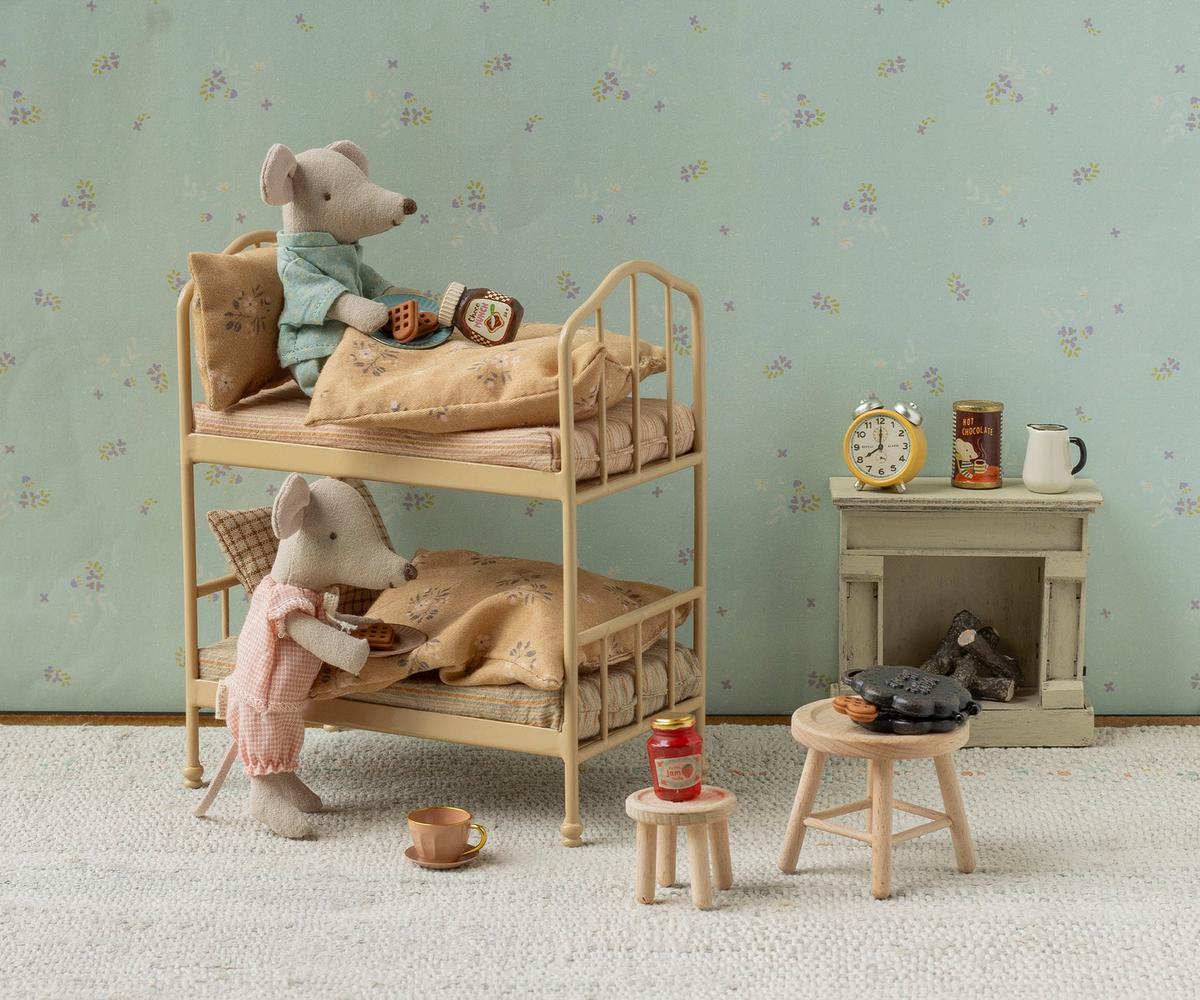 A miniature scene featuring two Maileg mice in a cozy room with floral wallpaper and a bunk bed, where one mouse sits on the top bunk and the other stands near a small wooden stool set up with the Maileg Waffle Set, including a waffle maker, cups, and jars.