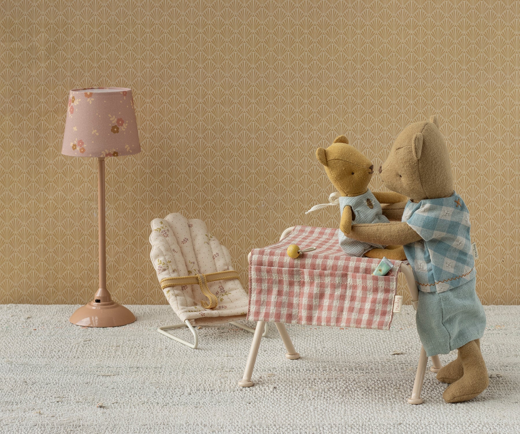 A miniature Maileg nursery scene with a floor lamp, a bouncy chair, and a small table covered in a pink gingham cloth where two toy teddy bears are seated, set against a warm beige patterned wall.