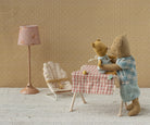 A miniature Maileg nursery scene with a floor lamp, a bouncy chair, and a small table covered in a pink gingham cloth where two toy teddy bears are seated, set against a warm beige patterned wall.