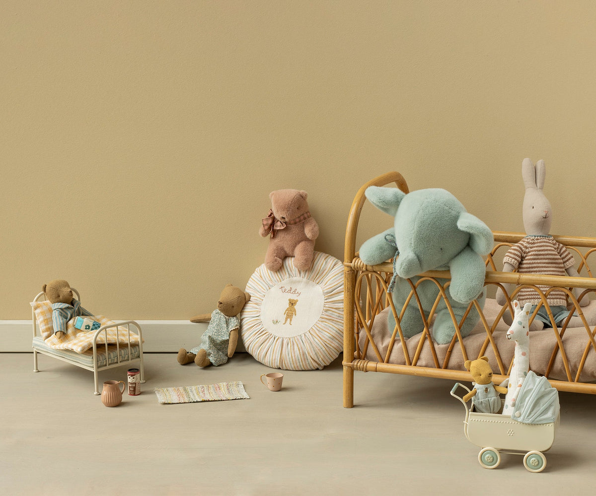 A styled collection of soft plush toys arranged in a nursery-like scene with a rattan crib, miniature beds, pillows, and a small toy stroller against a warm beige wall and floor.