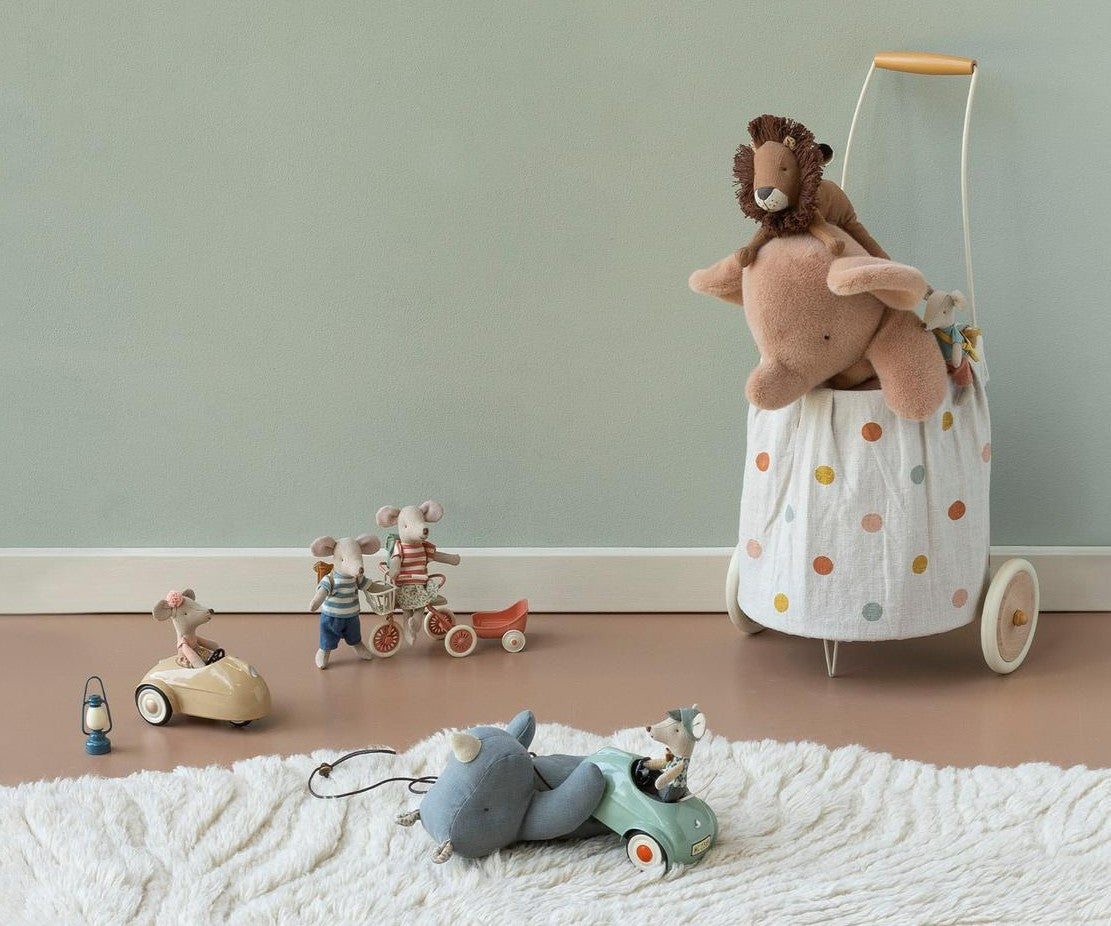 A styled collection of soft, vintage-style children's toys on a muted sage green wall. The set includes a large beige elephant plush toy, a brown lion, two small mouse figures on toy bicycles.