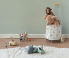A styled children's play scene with a white fabric toy cart covered in pastel polka dots, holding a plush stuffed animal. Nearby are small wooden toy cars, a tricycle, and other soft play items on a white textured rug against a pale green wall.