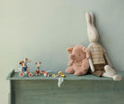 A styled collection of soft, vintage-style children's toys on a painted wooden shelf against a muted sage green wall. The set includes a large beige bunny plush toy in a striped shirt, a pink teddy bear, two small mouse figures on toy bicycles, and a small red toy car.