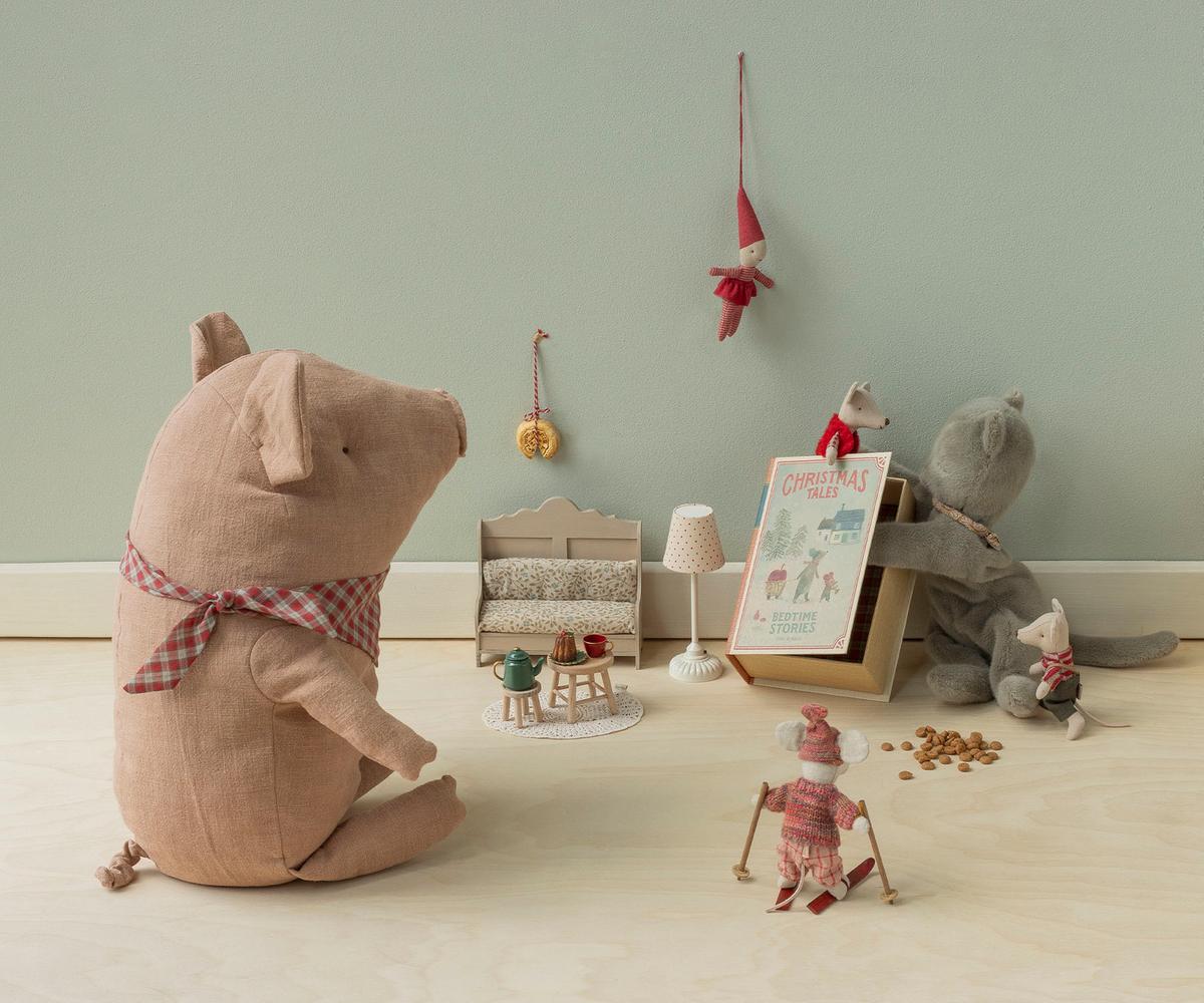A styled collection of soft fabric toys and miniature dollhouse accessories arranged on a light wood floor against a pale green wall. The set includes a large pinkish-brown pig plush wearing a red gingham bow, a gray mouse, a small red-and-white doll, a miniature lamp, a tiny table with a green tea set, and a hanging red elf ornament.