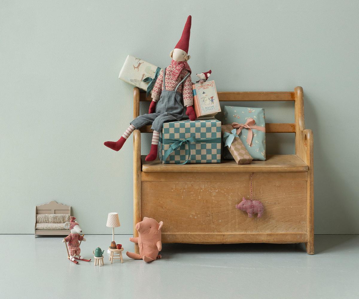 A styled nursery scene featuring a wooden toy chest bench with a gnome doll sitting on top, surrounded by wrapped gifts. In the foreground, a small plush pig and miniature dollhouse accessories sit on a light gray floor against a pale blue-gray wall.