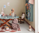 A cozy children's playroom scene featuring a large plush teddy bear wearing a plaid bow sitting on a patterned rug in the foreground, with a teal table, toy chairs, and various stuffed animals and toys in the background.