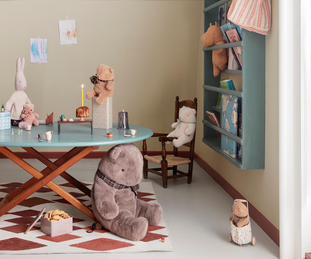 A larger plush teddy bear sits on a patterned rug in a children's room, while a smaller teddy bear rests near a wooden chair and table set. Additional toys, including stuffed animals and decorative elements, are displayed throughout the play area and on a wall shelf.
