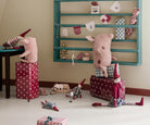 A toy room scene featuring Maileg mice characters: one sitting on a tall burgundy gift box at a small wooden table, another leaning against a large matching suitcase, and a toy dog lying near a miniature sofa bed against a teal wooden wall rack decorated with bunting.
