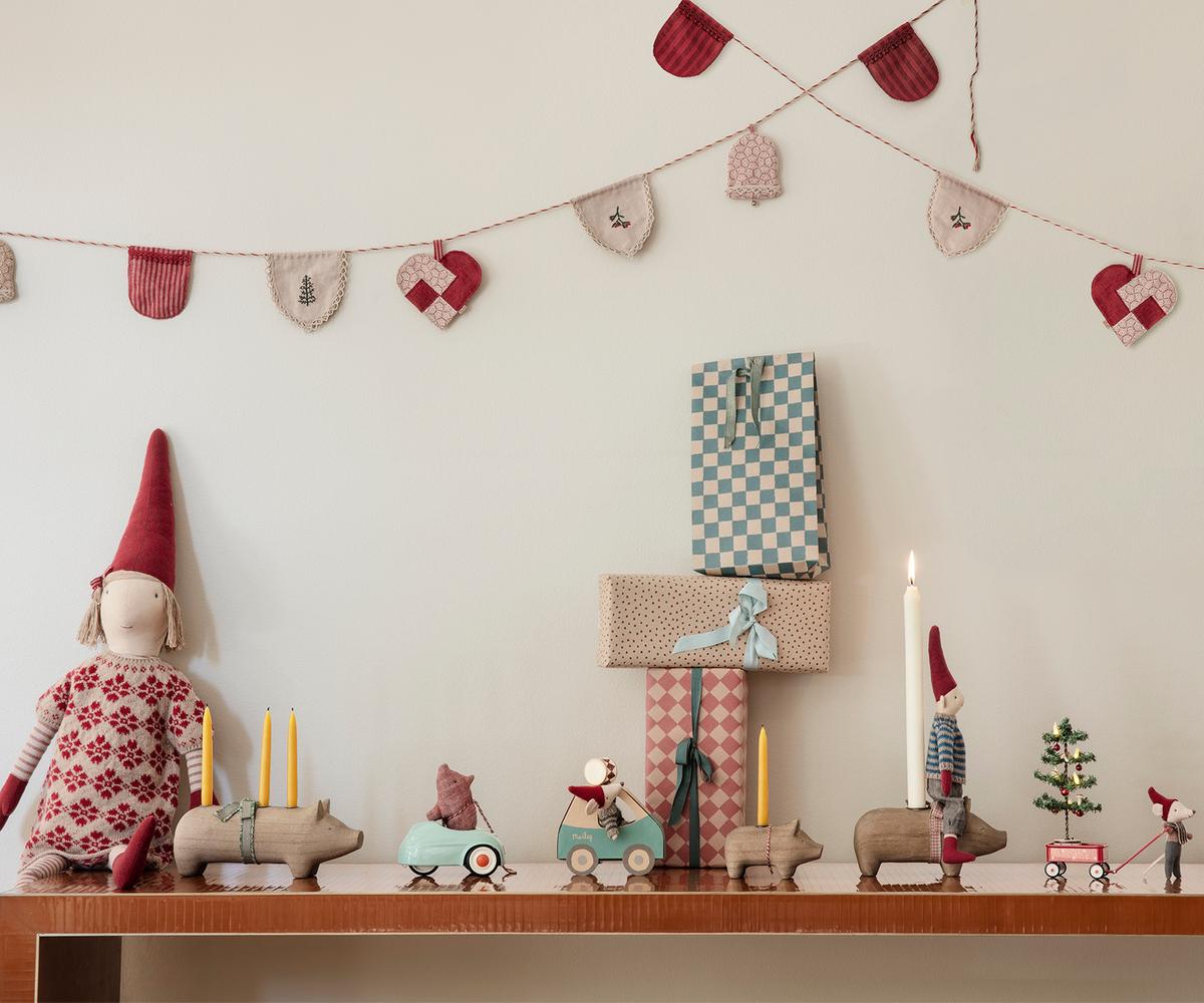 A festive holiday display featuring a mantelpiece decorated with wooden animal figures, miniature toy mice in toy cars, a lit candle, and wrapped gift boxes against a white wall strung with a felt heart and stocking garland.