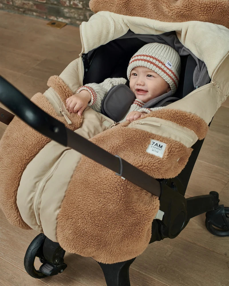 A baby wearing a striped beanie and pacifier is cozy inside an infant car seat outfitted with the Benji Applique Car Seat Cocoon by 7AM Enfant in oat with brown sherpa fleece trim.