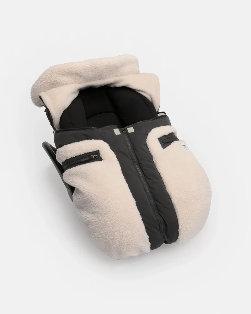 A baby footmuff in a warm cream fleece with black quilted panels and a black zipper, shown on a light gray background.