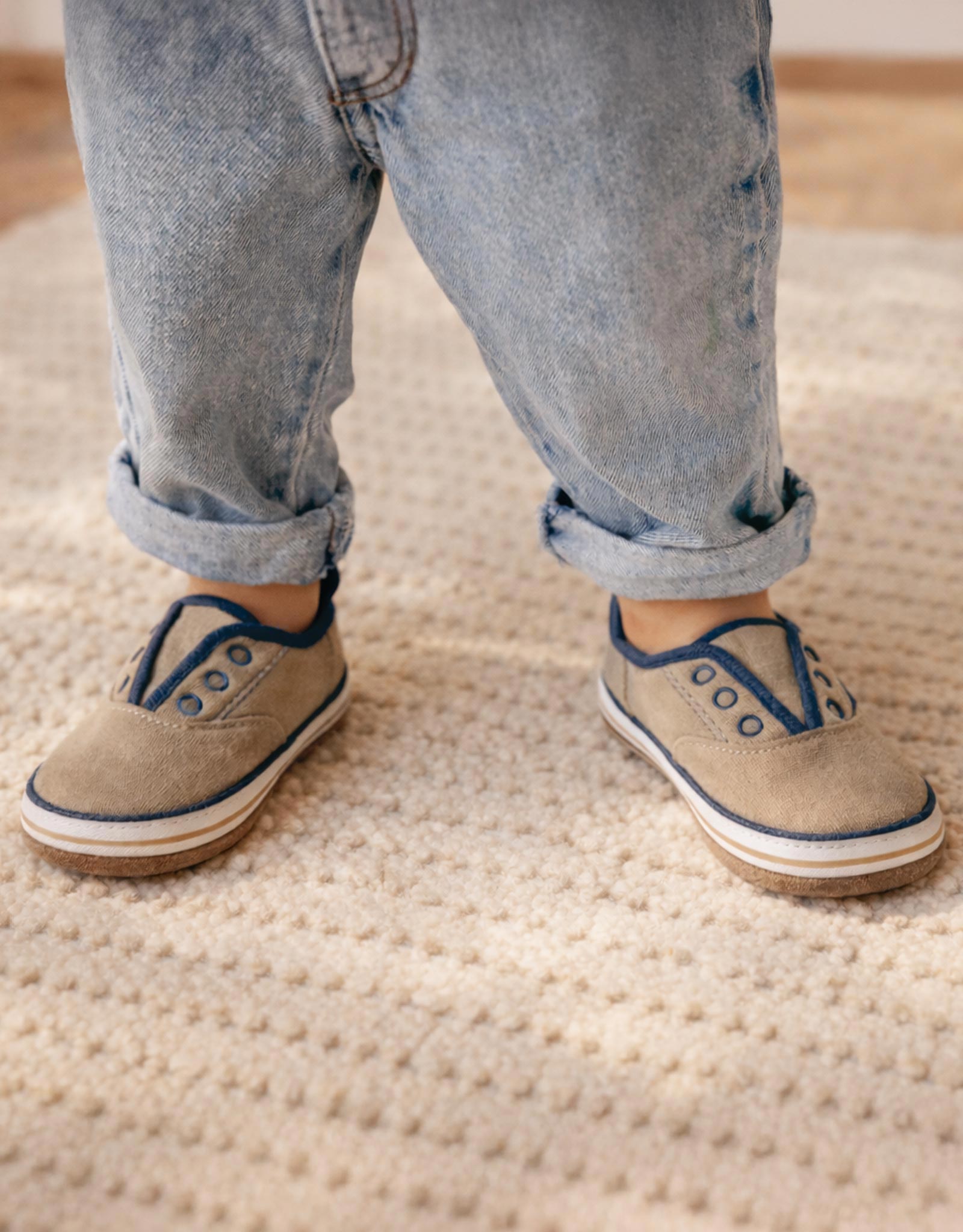 Person wearing beige shoes with blue laces and rolled-up jeans standing on a textured carpet.