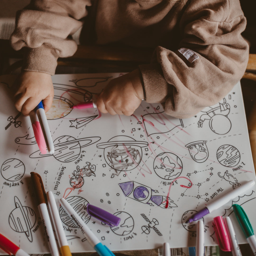 Child coloring a space-themed coloring book with markers on a wooden table.