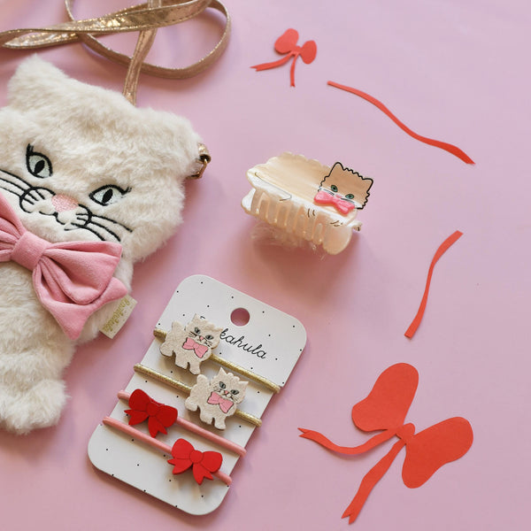 A styled flat-lay of cute cat-themed hair accessories on a pink background, including a white plush cat head with a pink bow, a cream claw clip with a cat face and bow, and a display card holding three small bow hair clips and two larger red bow hair ties.