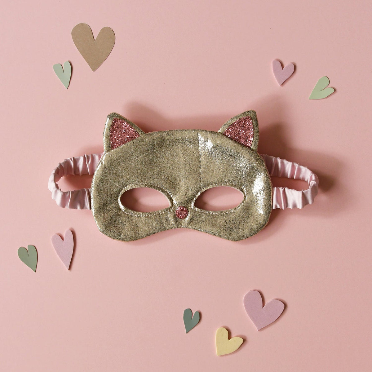 Gold cat mask with pink accents on a pink background with heart shapes