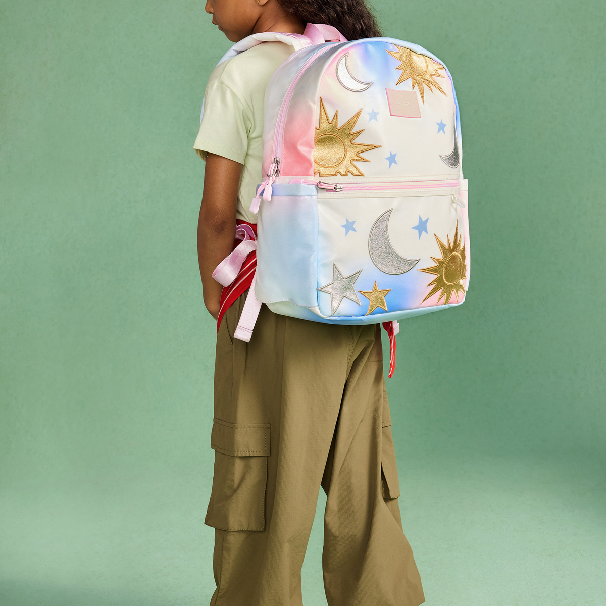 A child wearing a pastel rainbow backpack with gold sun and moon prints, shown from the back against a solid mint green background.