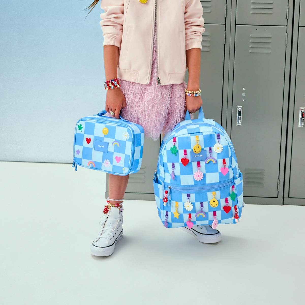 Person holding a blue checkered backpack and lunch bag with colorful designs in front of gray lockers.