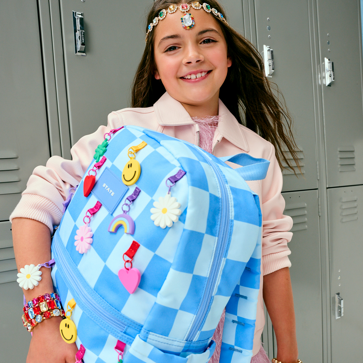 Girl holding a blue checkered backpack with colorful decorations in front of gray lockers.