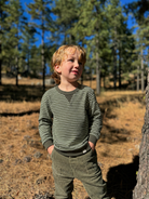 Young boy standing in a forested area with trees and dry grass in the background.