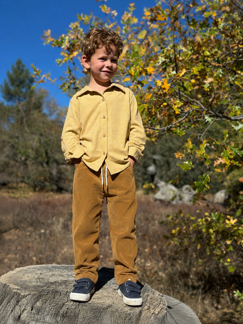 Child in a yellow shirt and brown pants standing on a log with trees in the background