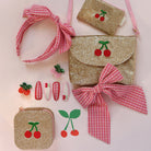 Set of gold glitter bags with cherry designs, pink headbands, and hair clips on a light pink background.