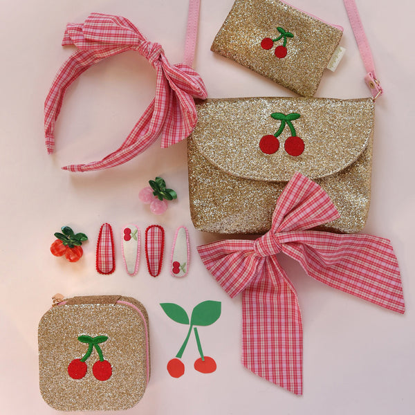 Set of gold glitter bags with cherry designs, pink headbands, and hair clips on a light pink background.