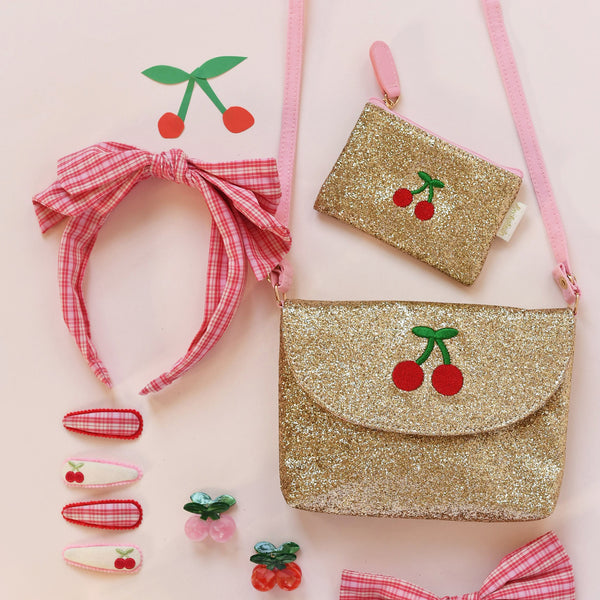 A coordinated cherry-themed accessory set on a pale pink background, including a glittery gold shoulder bag with a red-and-white gingham strap and cherry applique, a matching small pouch, a pink gingham bow headband, and several small hair clips and decorative cherry pieces.