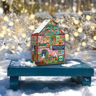 A colorful 100-piece Christmas-themed jigsaw puzzle box shaped like a house, displayed outdoors on a blue bench in the snow with blurred holiday lights in the background.