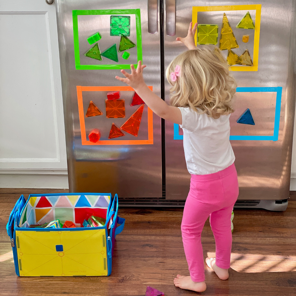 A young child standing in front of a stainless steel refrigerator, attaching colorful translucent magnetic tiles to colorful frames adhered to the fridge, with a blue toy storage bin filled with more tiles on the floor nearby.