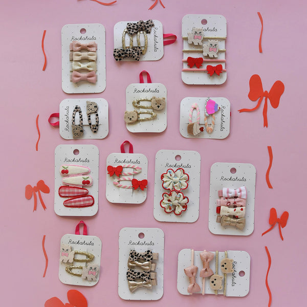 Twelve small hair clips displayed on white hanging cards against a pink background with red ribbon decorations. The clips include bows, flowers, teddy bears, hearts, and animal motifs in pastel pink, cream, red, beige, and black-and-white patterns.
